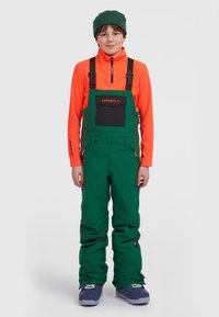 Groene sneeuwoveralls met verstelbare bandjes, een voorzak en oranje accenten, gecombineerd met een feloranje rits-jack en een groene muts.