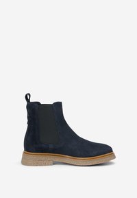 Botte Chelsea en daim bleu marine avec panneaux latéraux élastiques noirs, semelle en caoutchouc couleur caramel et languette de tirage au talon, présentée sur un fond blanc.