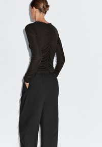 Zwarte top met lange mouwen en geplooide achterkant, met een gegolfde halslijn en zachte textuur, gecombineerd met loszittende zwarte broek.
