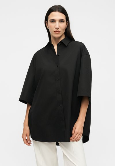 Γυναίκα με ίσια σκουρα μαλλιά, φοράει υπερμεγέθη μαύρο πουκάμισο με κουμπιά και άσπρο παντελόνι, στέκεται μπροστά σε απλό φωτεινό φόντο.