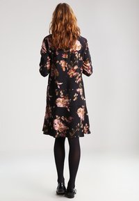 Schwarzes Blumenmusterkleid mit pinken und goldenen Mustern, langen Ärmeln, knielanger Länge und lockerer Passform. Getragen mit schwarzen Strumpfhosen und flachen Schuhen.