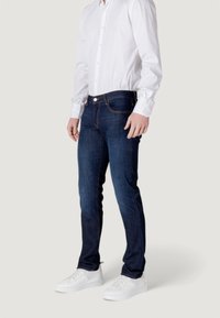 Jeans ajustados de color azul oscuro con una textura suave, diseño de cinco bolsillos y costuras en contraste, combinados con una camisa blanca de botones y zapatillas blancas.