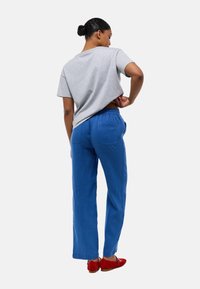 T-shirt gris à manches courtes, pantalon en lin bleu et chaussures rouges. La vue arrière montre une coupe décontractée avec une taille élastique et des poches à rabat.