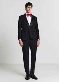 Frack nero con collo sciallato in raso, bottoni neri e pantaloni coordinati. Camicia bianca e papillon rosso, abbinati a scarpe nere lucide.