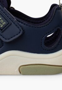 Gros plan d'une sandale pour enfant bleu marine avec sangle à velcro, col rembourré, semelle blanche et semelle texturée vert clair.