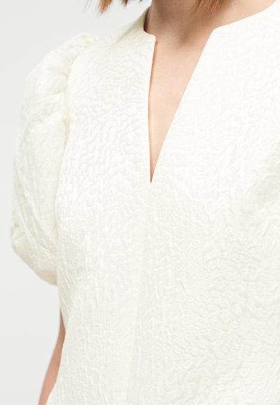 Blusa branca texturizada com mangas curtas bufantes e decote em V profundo. O tecido apresenta um padrão subtil em relevo.