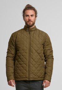 Vihreä tikattu takki, jossa on pystykaulus, vetoketju ja napit. Takissa on sivutaskut ja teksturoidut ompelukuvio.