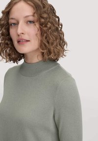 Hellgrüner, figurbetonter Pullover mit hohem, geripptem Kragen, weicher Textur und glattem Strickmuster, vor einem neutralen Hintergrund präsentiert.