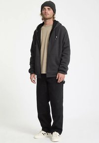 Zwarte hoodie met rits, gedragen over een beige sweatshirt, gecombineerd met zwarte cargobroek en witte sneakers. Afgewerkt met een casual gebreide muts.