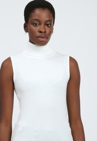 Maglione senza maniche bianco in una morbida trama lavorata a maglia. Presenta un collo alto e una silhouette aderente.