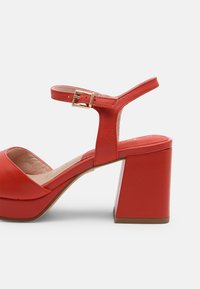 Sandale à talons hauts en cuir rouge avec un bout arrondi, une bride de cheville avec boucle dorée et un talon carré épais. Texture lisse sur toute la surface.