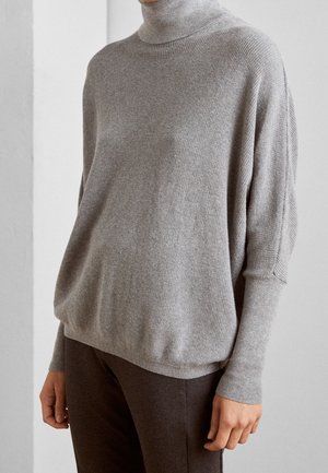 Osoba nosící volný světle šedý rolákový svetr s dlouhými přiléhavými rukávy a tmavě hnědými kalhotami, stojící uvnitř proti jednoduché stěně.