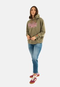 Olijfgroene hoodie met roze tekst, relaxed fit en een grote capuchon. gecombineerd met lichtblauwe spijkerbroek en rode sneakers met witte accenten.