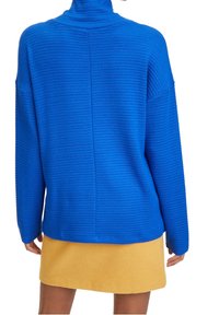 Suéter azul acanalado con cuello alto y hombros caídos, combinado con una falda amarilla, que muestra una tela con textura y un ajuste relajado.