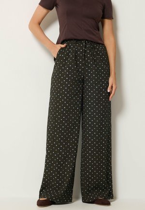 Donna con pantaloni larghi scuri con pois azzurri chiari e una maglietta scura a maniche corte, in piedi con una mano in tasca.