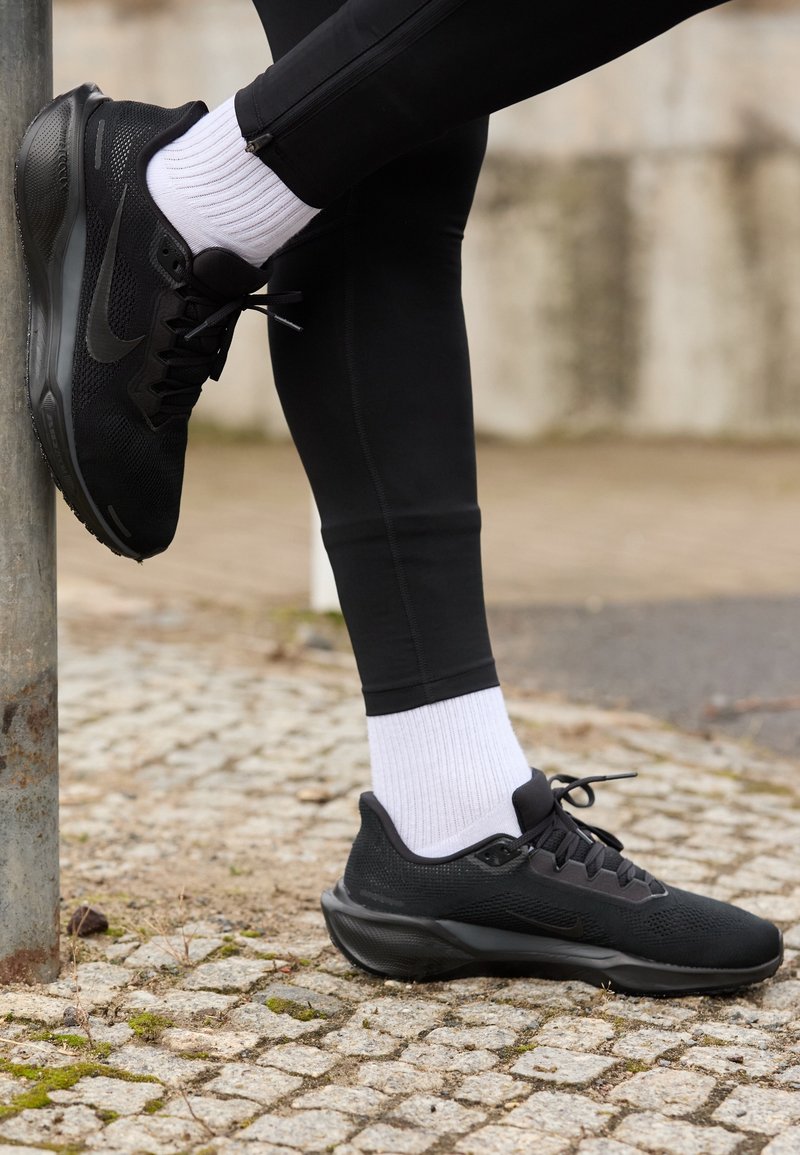 Personne portant des baskets noires et des chaussettes blanches, étirant une jambe contre un poteau métallique sur un pavé en pierres.