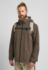 Bruine waterdichte jas met een ritssluiting aan de voorkant, elastische manchetten en een verstelbare capuchon. Gekozen in combinatie met een beige rugzak met een borstriem.