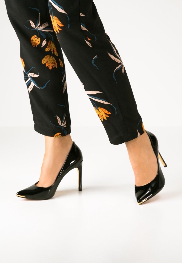 Pieds portant des escarpins en cuir verni noir et un pantalon noir avec des imprimés floraux orange sur fond blanc.