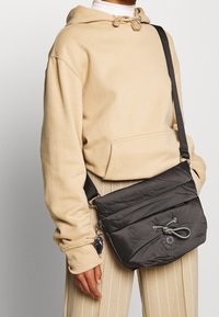 Czarna pikowana torebka crossbody z zamknięciem na sznurki, naszywką z logo i regulowanym paskiem, w zestawie z beżową bluzą z kapturem i paskowanymi spodniami.