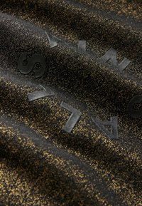 Tela negra con un brillo dorado metálico, que presenta letras en negro mate ligeramente elevadas. La textura parece suave y texturizada.