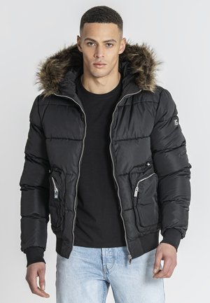 Mann trägt eine schwarze Steppjacke mit pelzbesetzter Kapuze, schwarzen Hemd und hellblauen Jeans, steht vor einem schlichten Hintergrund.
