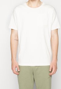 Bílá bavlněná tričko s kapsičkou na hrudi, kulatým výstřihem a krátkými rukávy. Měkká textura; obsahuje decentní branding na kapsičce.