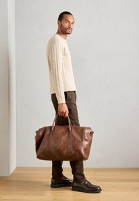 Man standing indoors wearing beige cable knit sweater, brown pants and shoes, holding large brown leather duffel bag by side.