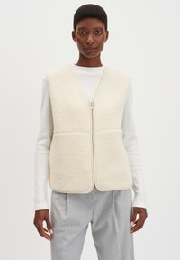 Einen cremefarbenen Fleece-Weste mit Reißverschluss, die eine weiche Textur und eine taillierte Form hat, kombiniert mit einem weißen Langarmoberteil und grauen Hosen.