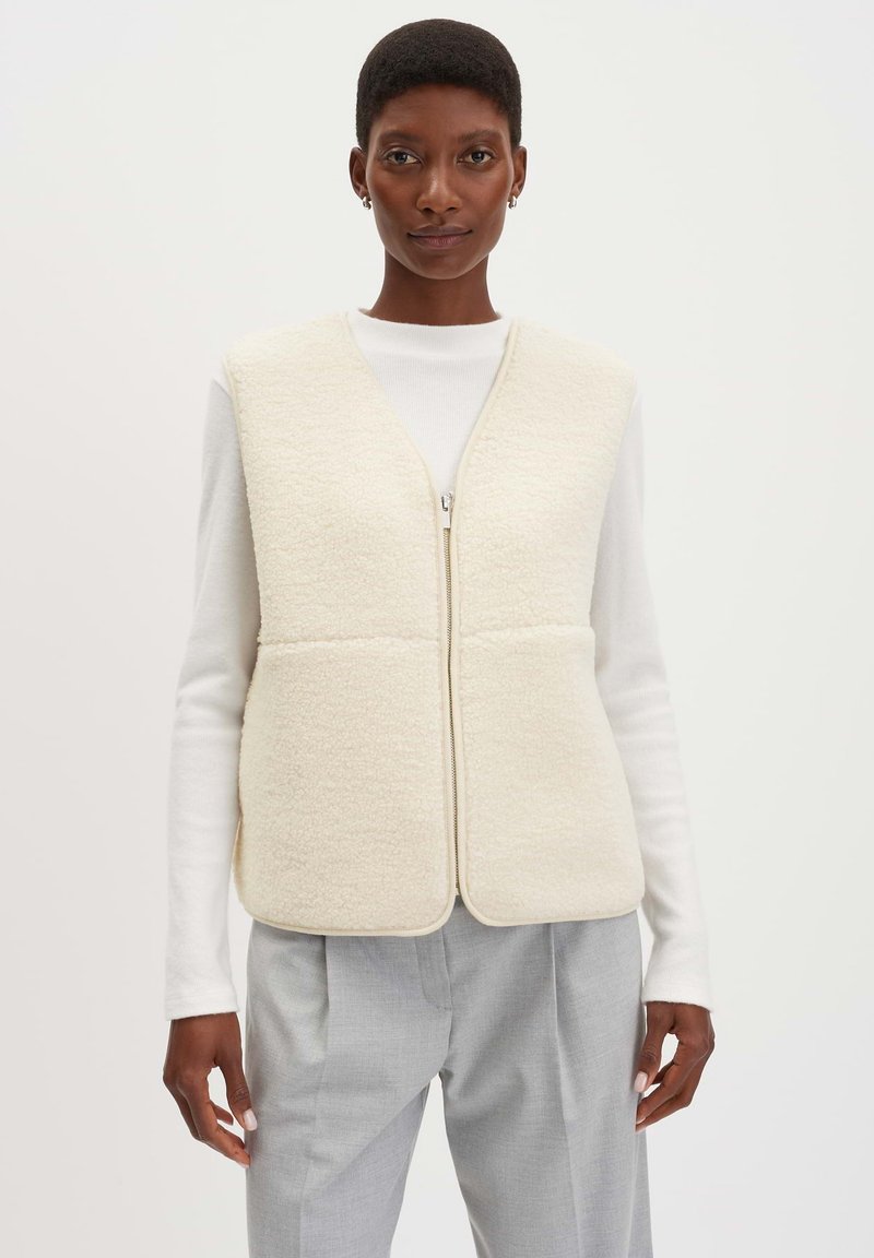 Einen cremefarbenen Fleece-Weste mit Reißverschluss, die eine weiche Textur und eine taillierte Form hat, kombiniert mit einem weißen Langarmoberteil und grauen Hosen.