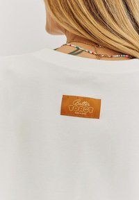 Bílá mikina s kulatým výstřihem a obdélníkovou oranžovou nálepkou se textem "Butter Toast MORNINGS" na zádech, texturovaná tkanina, ležérní design.