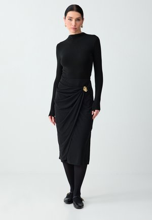 Donna che indossa una maglia nera aderente a maniche lunghe e una gonna midi nera drappeggiata con spilla dorata, calze nere e scarpe nere, in piedi su sfondo bianco.