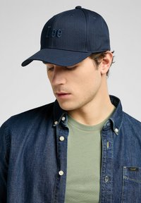 Marine pet met geborduurd "Lee" logo, gemaakt van gladde stof. Gecombineerd met een spijkerhemd en een lichtgroene t-shirt eronder.