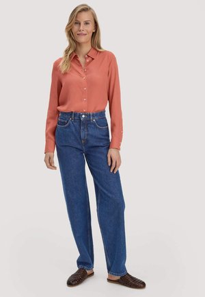 Vrouw staand, gekleed in een roestkleurige blouse met knopen, blauwe high-waisted jeans en donkerbruine instapschoenen tegen een effen achtergrond.