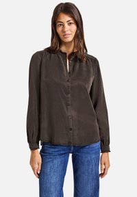 Blouse verte foncée à manches longues, avec un boutonnage sur le devant, des détails plissés aux épaules et des poignets élastiqués. Tissu doux et lisse.