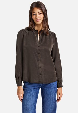 Blouse verte foncée à manches longues, avec un boutonnage sur le devant, des détails plissés aux épaules et des poignets élastiqués. Tissu doux et lisse.