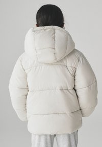 Krem boje bunda s kapuljačom velikih dimenzija, s kavljastim dijelovima i mekim, teksturiranim materijalom. Topli i široki dizajn.