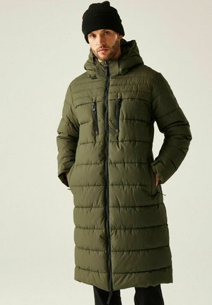 Mann trägt langen olivgrünen gepolsterten Wintermantel mit Reißverschlusstaschen, schwarze Mütze, steht mit Händen in den Taschen vor neutralem Hintergrund.