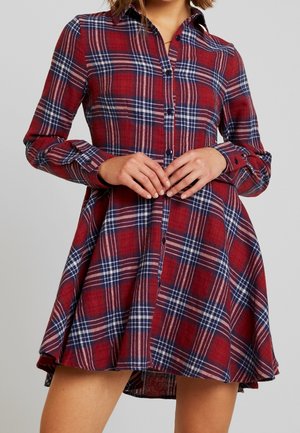 Mujer con un vestido abotonado a cuadros rojo, azul y blanco, con mangas largas y falda acampanada, juntando las manos a la altura de la cintura.