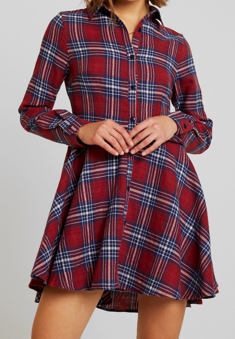 Femme portant une robe chemise à carreaux rouge, bleu et blanc, avec des manches longues et une jupe évasée, les mains jointes au niveau de la taille.