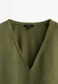 Haut en lin vert olive à col en V avec des bords bruts autour de l'encolure et une étiquette noire à l'intérieur du col.