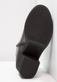 Black leather shoe with a textured rubber sole, featuring a block heel and a decorative strap detail on the side.