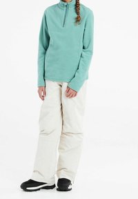 Pullover in pile turchese chiaro con mezza zip, abbinato a pantaloni da neve color crema. Stivali da neve neri completano l'outfit.