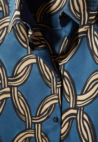 Tissu bleu avec un motif de chaînes entrelacées beige et noir sur un col de chemise boutonné et le devant.