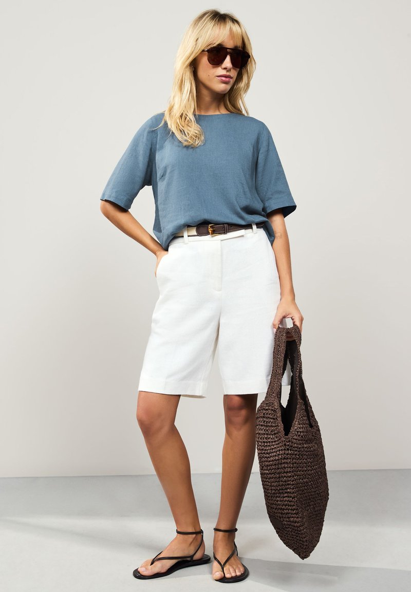 Blue linen short-sleeve top, white tailored shorts, black flat sandals, and a brown woven tote bag. Plain textures, neutral colors.