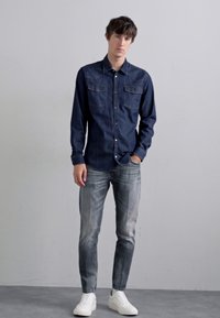 Camicia di jeans blu scuro con design a bottoni, dotata di due tasche sul petto e un colletto, abbinata a jeans grigi sbiaditi e sneaker bianche.