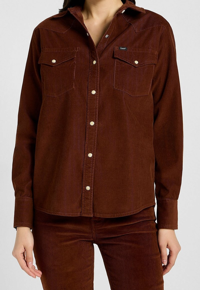 Femme portant une chemise marron en velours côtelé à boutons avec deux poches poitrine devant, et un pantalon assorti en velours côtelé marron.