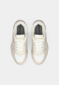Pár béžových a bielych tenisiek Reebok s bielymi šnúrkami, pohľad zhora na jednoduchom bielom pozadí.