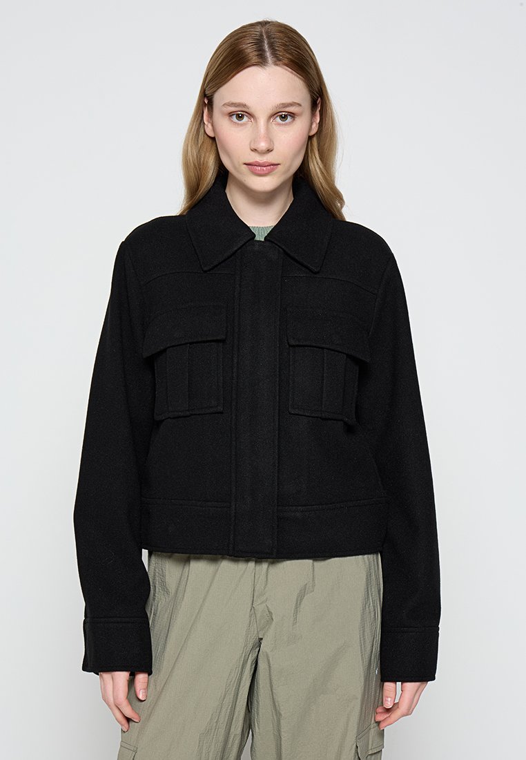 Veste noire courte en tissu texturé, avec fermeture éclair à l'avant, grandes poches poitrine et col pointu.