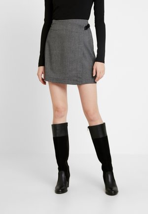 Femme portant une mini-jupe grise avec une boucle latérale, un haut noir à manches longues et des bottes noires montantes, debout sur un fond blanc.