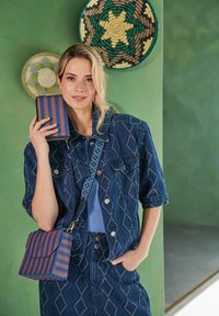 Denimjack met diamanten stiksels, gecombineerd met een blauw geribbeld topje en een gestreepte blauw-gele schoudertas. Groene achtergronddecoratie.
